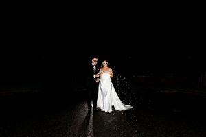 Bride and groom stand together at night in the rain, illuminated against a dark background in elegant wedding attire.