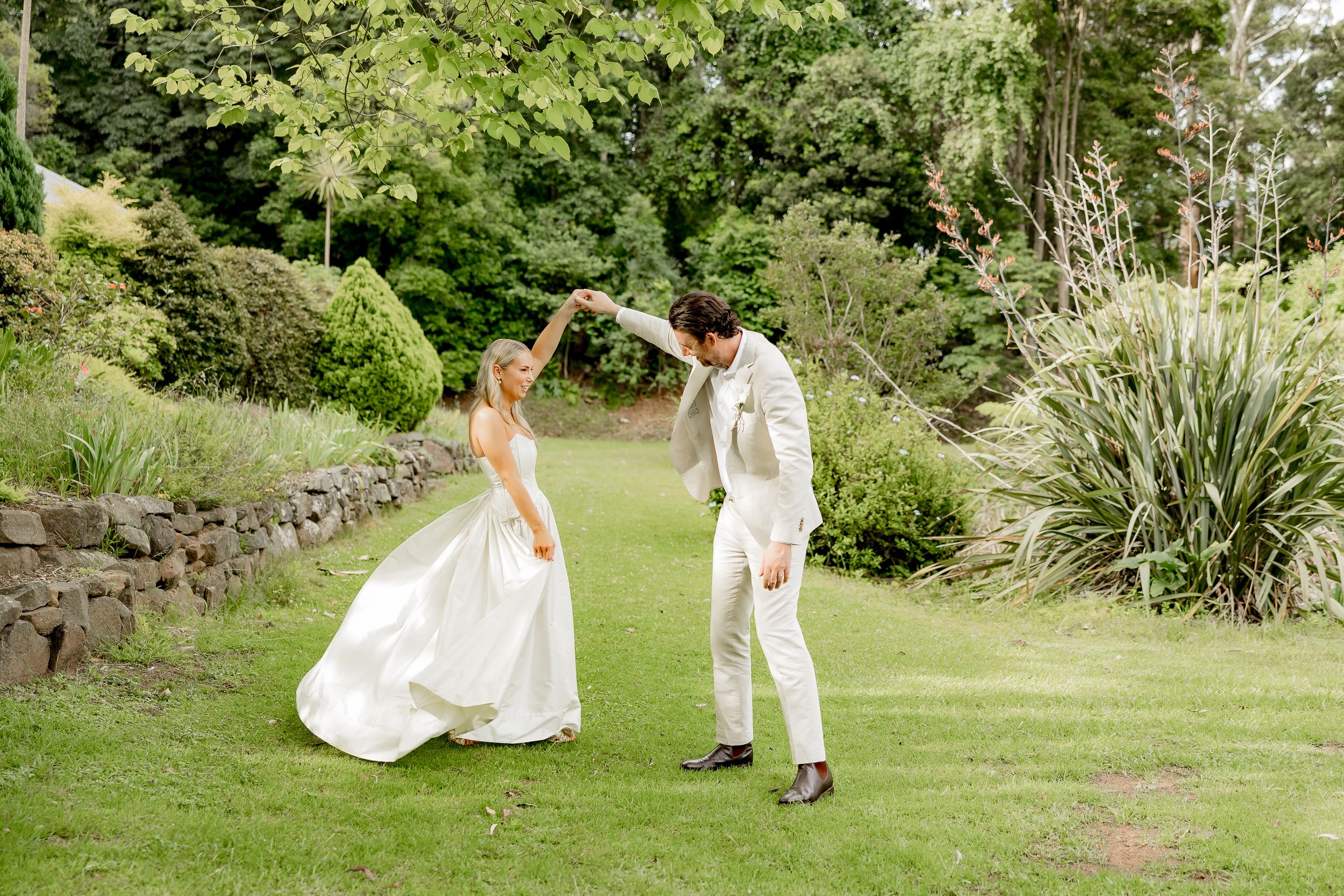 Two people in white outfits dancing together on a lush green lawn surrounded by garden landscaping.