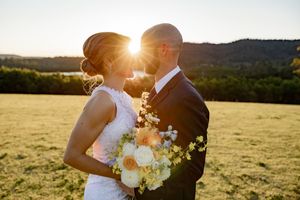 A romantic sunset wedding photo in a golden field with a bouquet of white flowers and sunlight glowing behind the couple.