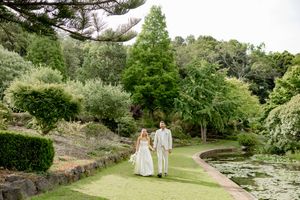 A romantic couple walks hand in hand through a lush garden path surrounded by blooming flowers and greenery.