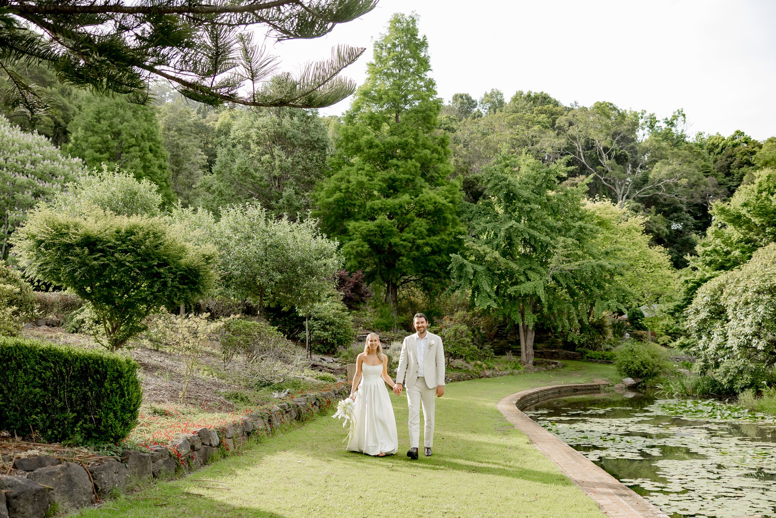 A romantic couple walks hand in hand through a lush garden path surrounded by blooming flowers and greenery.
