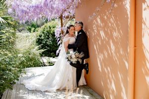 A romantic wedding moment captured under blooming wisteria against a peach-colored wall in a garden setting.