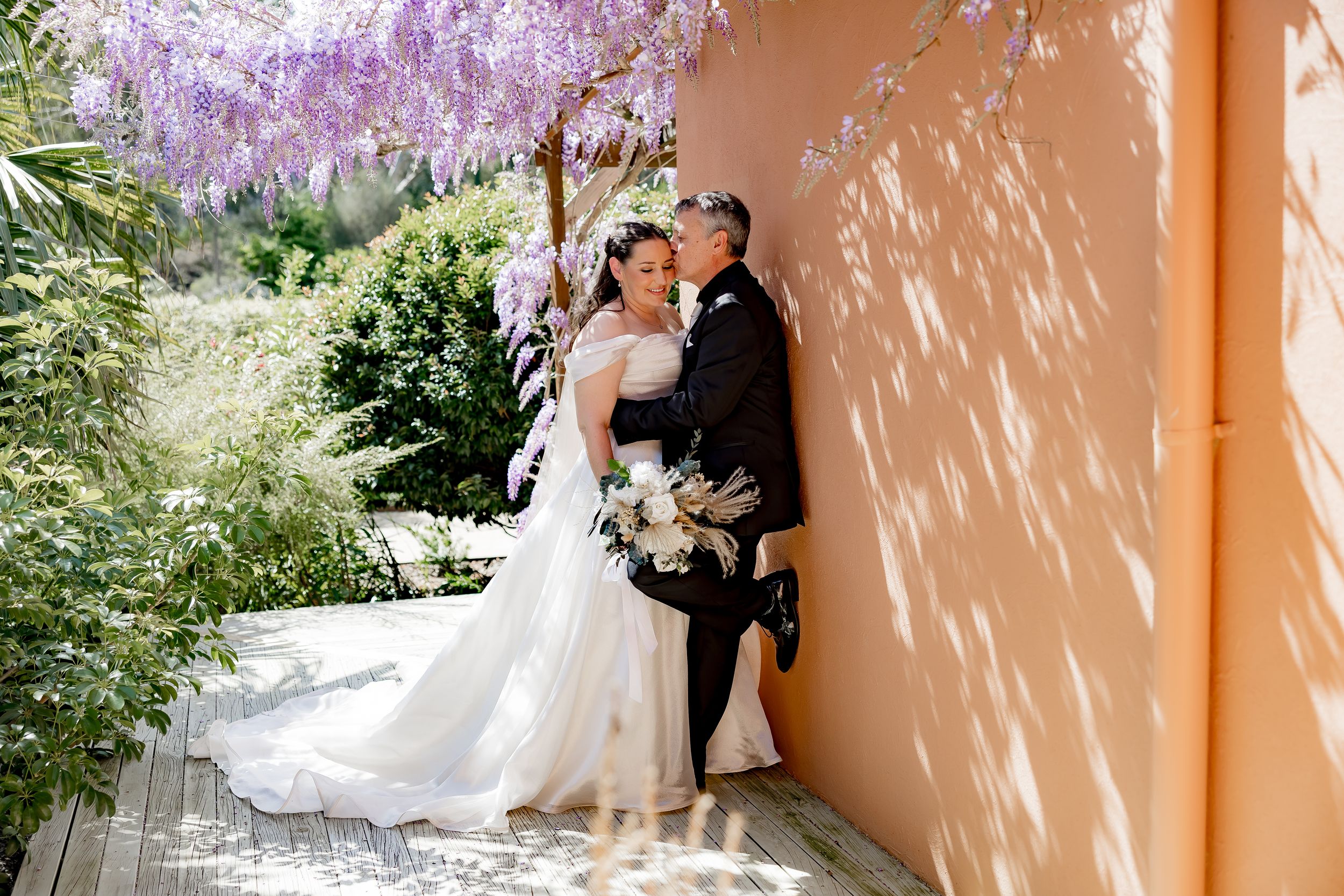 A romantic wedding moment captured under blooming wisteria against a peach-colored wall in a garden setting.