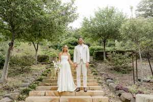 A couple in white attire poses together on rustic stone steps surrounded by lush garden landscaping.