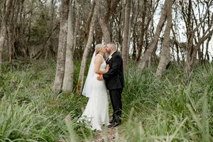 A couple in wedding attire shares a romantic moment among tall trees in a wooded forest setting.