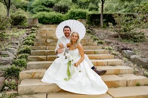 Wedding couple shares an intimate moment on outdoor stone steps with white parasol and floral details.