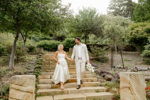 Two figures in white attire walk together through a lush garden pathway on a sunny afternoon.