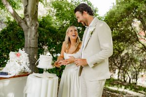 A couple in light-colored wedding attire share a joyful moment together at their outdoor reception by the cake table.