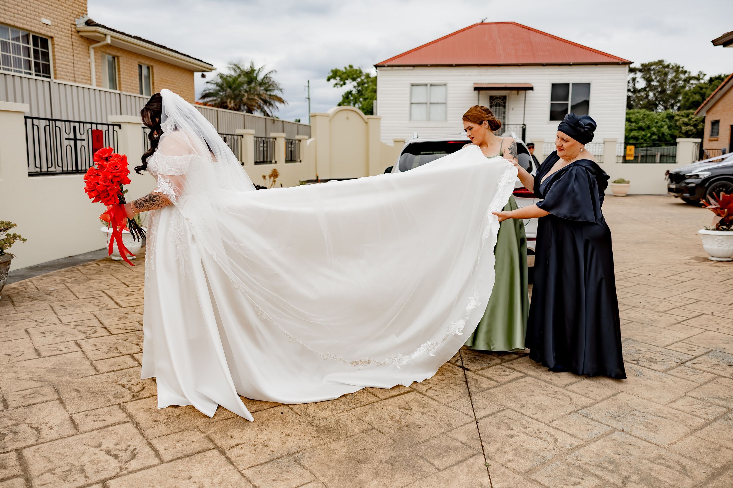 People in formal attire help arrange a flowing white wedding dress train on a stone patio outside.