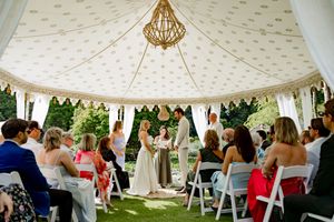 Outdoor wedding ceremony under an elegant white tent with chandelier and guests seated on white chairs.