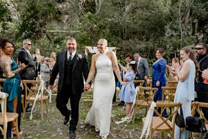 Wedding guests gather outdoors at a rustic venue with wooden benches and natural greenery for a ceremony.