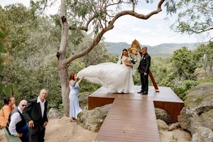 A wedding ceremony takes place on a wooden deck overlooking scenic mountain views with eucalyptus trees framing the scene.