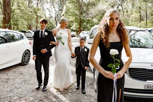 A wedding party walks through a wooded area wearing formal black and white attire while holding bouquets.