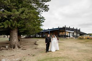 A couple in wedding attire stands near a large tree with an industrial structure in the background at an outdoor venue.