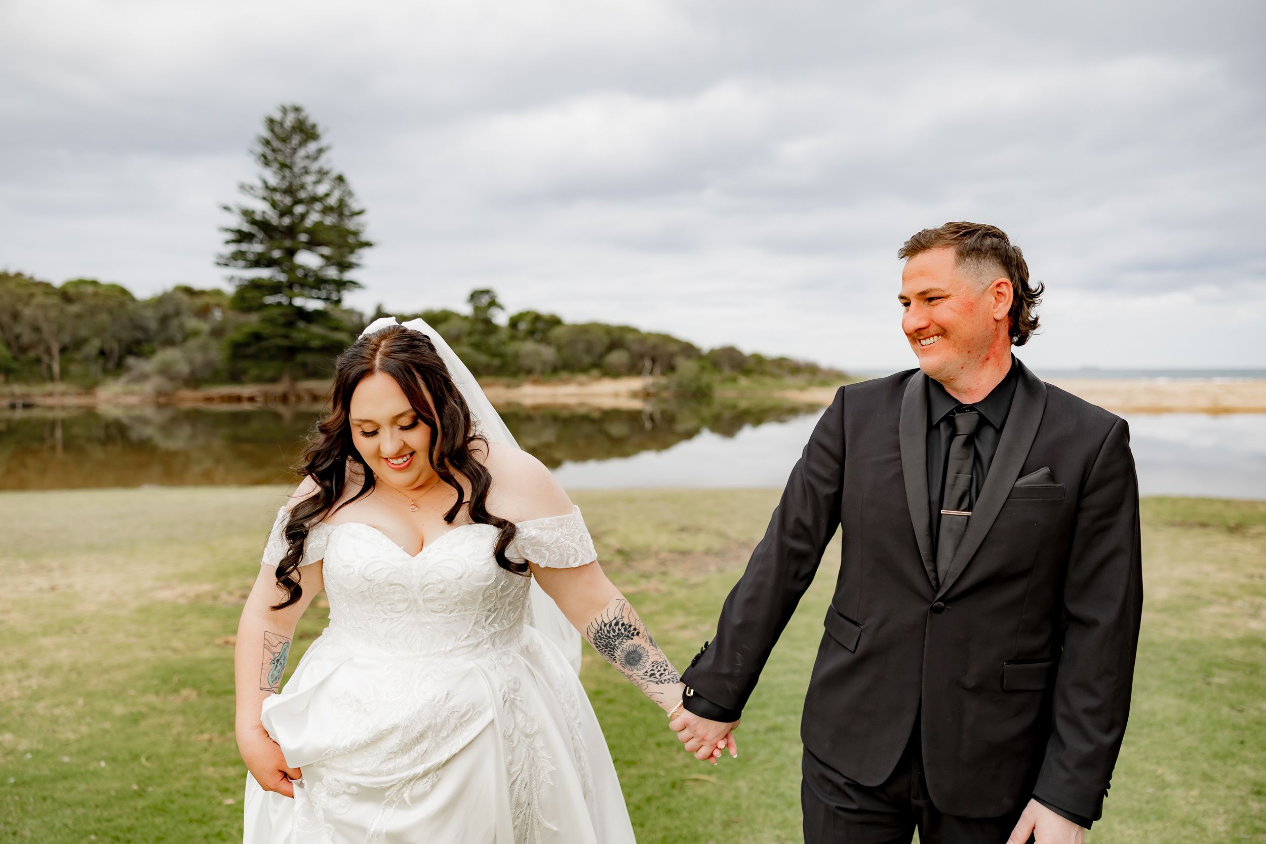 A couple in wedding attire holds hands and walks together across a scenic coastal landscape under cloudy skies.