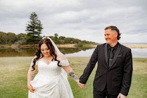 A couple in wedding attire holds hands and walks together across a scenic coastal landscape under cloudy skies.