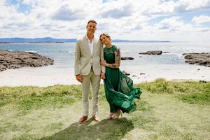 A couple in formal attire poses together on a scenic coastal beach with blue water and cloudy skies in the background.