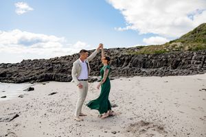 A couple walks along a scenic rocky beach coastline with dramatic cliffs and white sand on a sunny day.