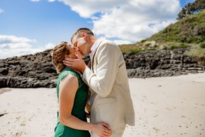 A couple in formal attire sharing romantic moments on a scenic beach with rocky cliffs in the background.