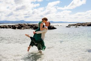 A couple embraces in emerald green dress on a pristine beach with rocky outcrops and turquoise waters in the background.
