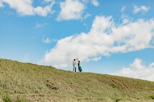 Two people stand together on a grassy hilltop overlooking a bright blue sky with puffy white clouds.