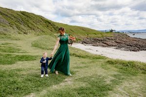 A couple in formal attire poses on a grassy coastal hillside with ocean views in the background.