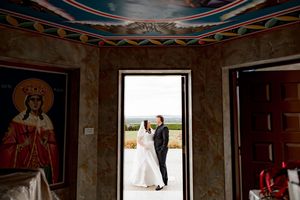 A couple stands in a church doorway framed by religious artwork and decorative walls, with natural light streaming in.