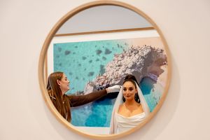 A bride getting her veil adjusted by a stylist, reflected in a round wooden mirror against a white wall.