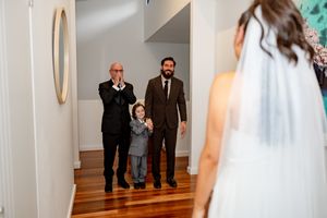 Emotional first look moment as bride reveals herself to groom, father, and child in hallway on wedding day.