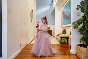 Young girl in pink tulle gown holding red flower toy walks through modern home hallway with timber floors.