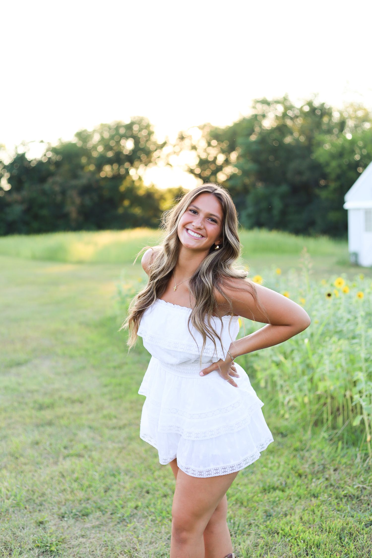 A girl in a white strapless dress smiling in a field near a barn.