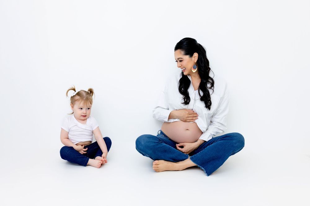 A pregnant woman in white sits cross-legged beside a toddler with pigtails on a clean white backdrop.