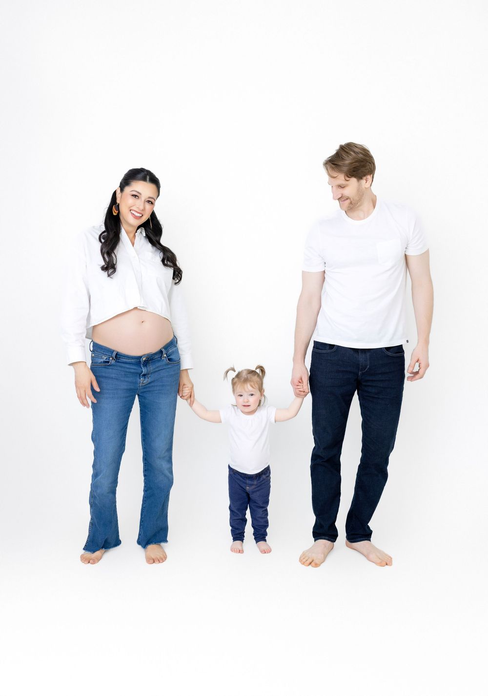 Parents and child holding hands while walking barefoot against white background in casual outfits.