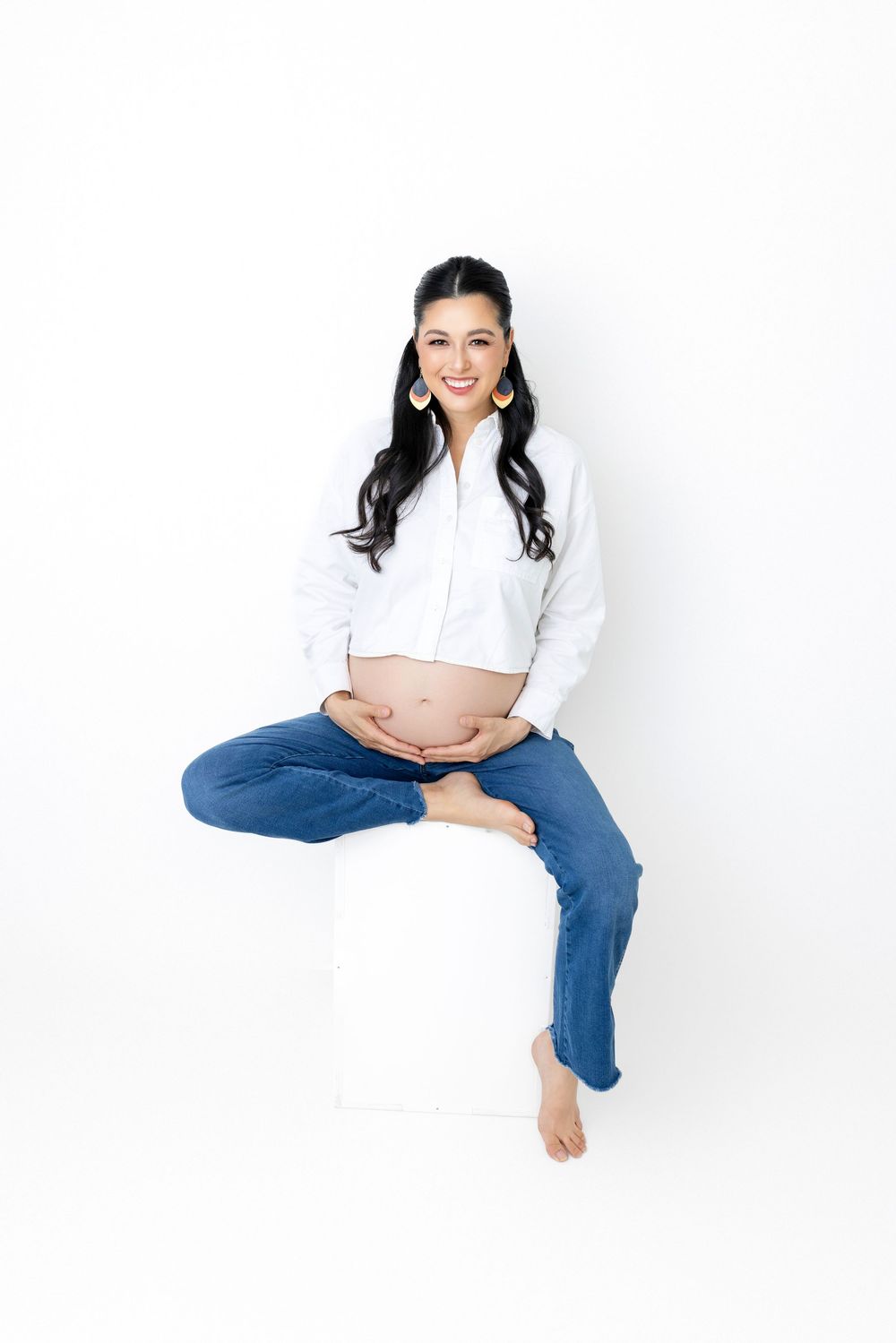Expectant mother in white cropped shirt and jeans sits casually against white background showing pregnant belly.