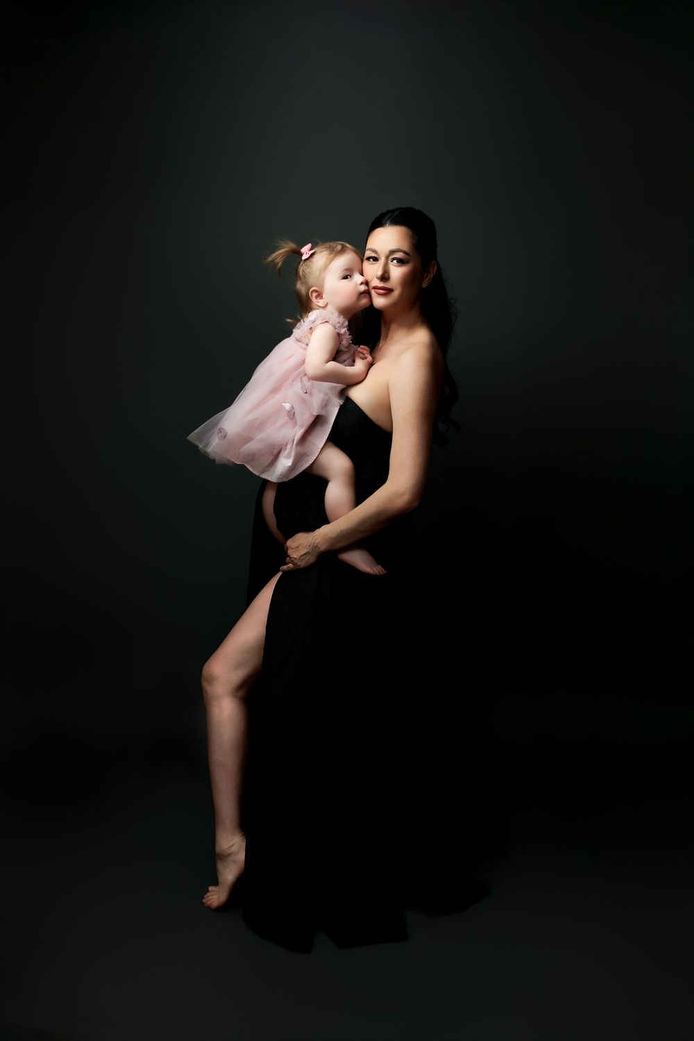 A tender portrait of a parent and child dancing together against a dark background in a flowing dress and ballet attire.