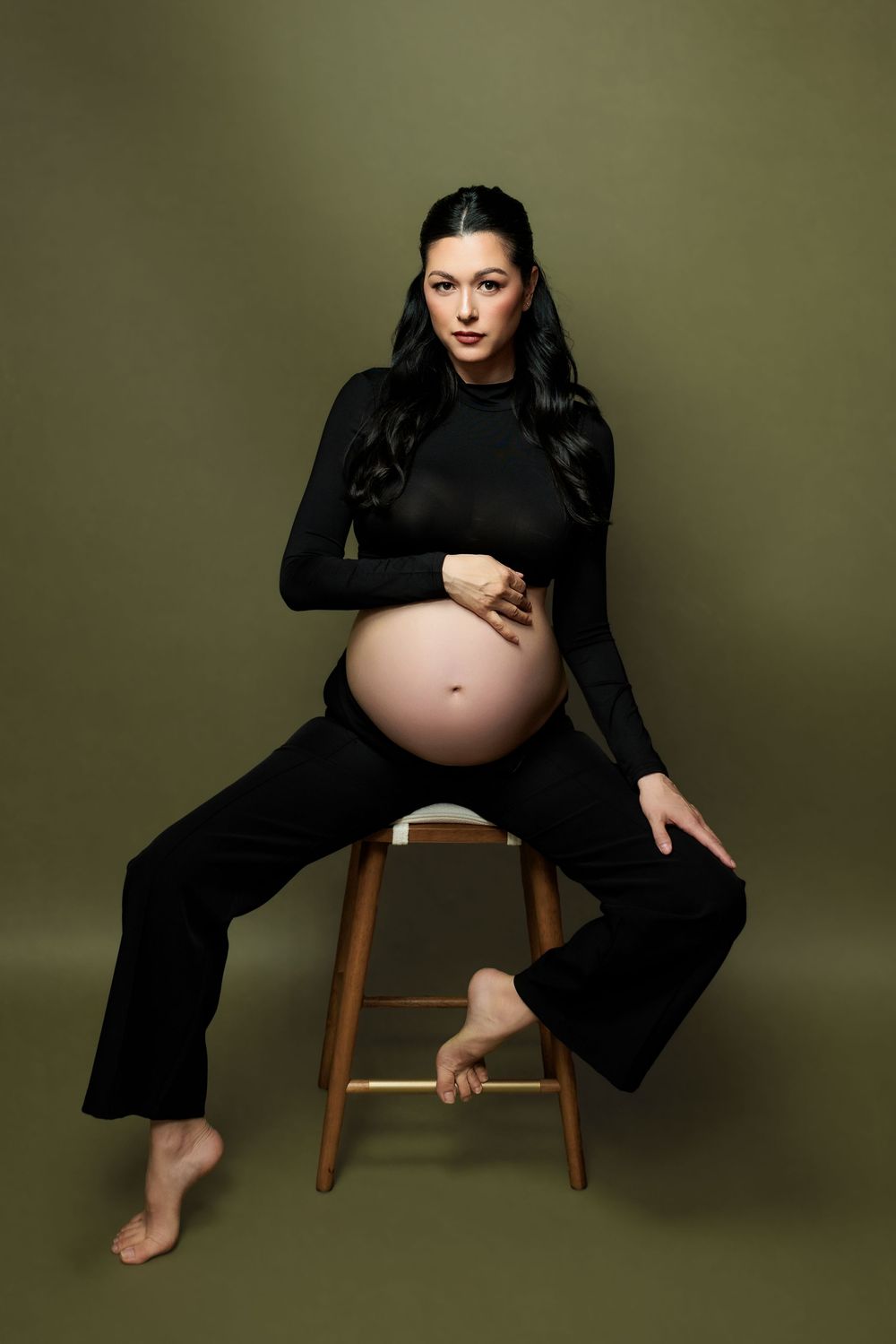 A woman in a black dress sits on a wooden chair against a dark backdrop highlighting her pregnant silhouette.