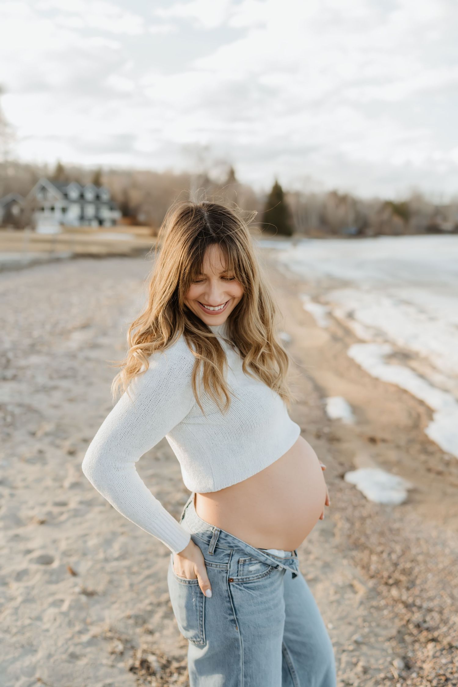 Expectant mother in white sweater and jeans poses on beach during winter.