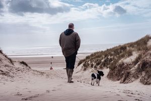 Man walking dog on sandy beach dunes with ocean view and cloudy sky in background.