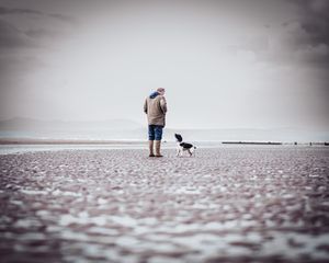 A man and his small dog stand alone on a vast, desolate beach under an overcast sky.