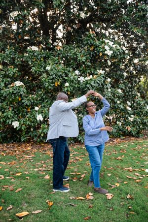 Playful couple bending forward and laughing in garden with fallen leaves scattered on grass.