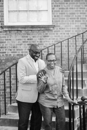 Black and white photo of couple laughing together by brick building with metal railing.