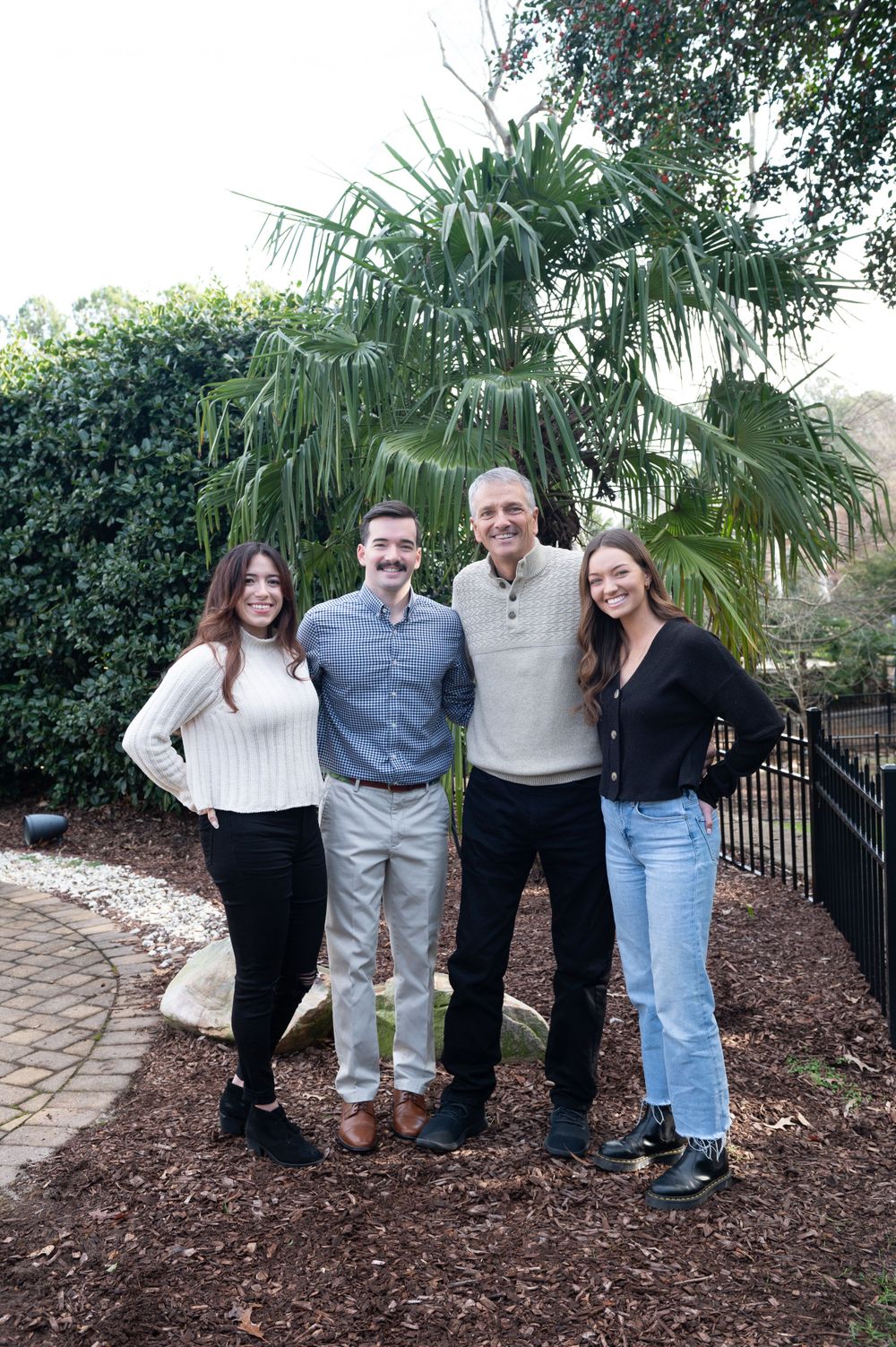 Group of people posing together in casual attire against landscaped garden with palm trees.