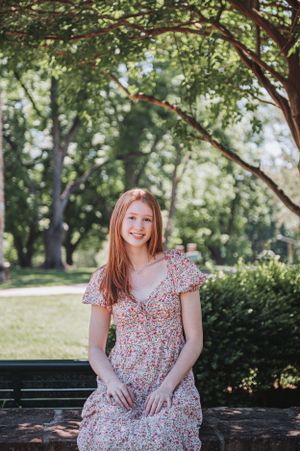 Woman in floral dress sitting on stone wall surrounded by lush green trees in outdoor park setting.