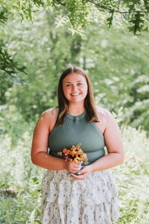 Woman in olive top and floral skirt holding flowers in sunlit garden.