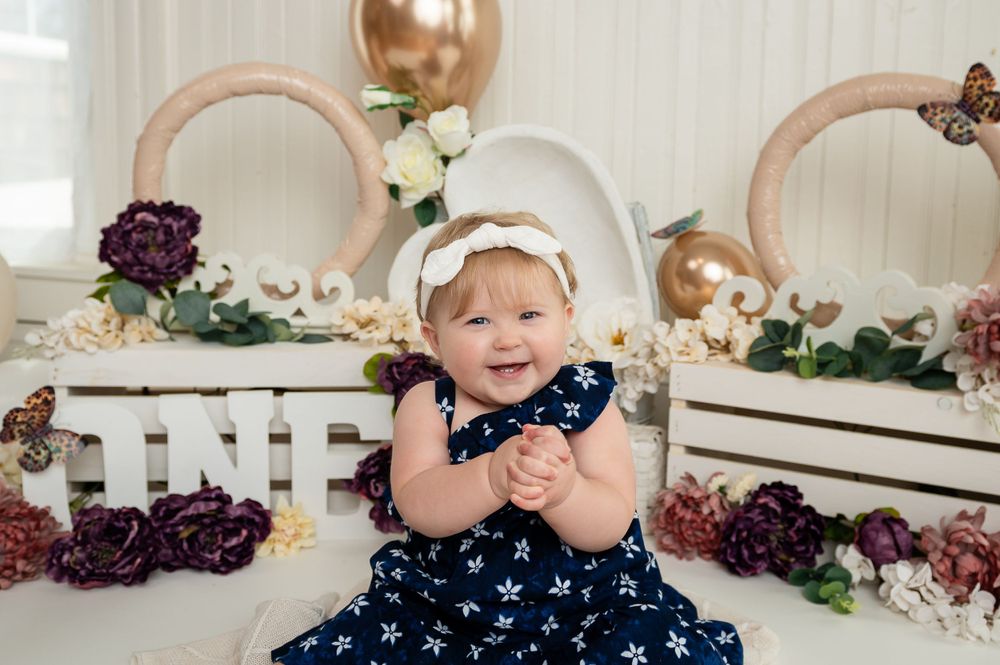 Baby in navy star dress poses during cake smash photoshoot with white and rose gold decorations.