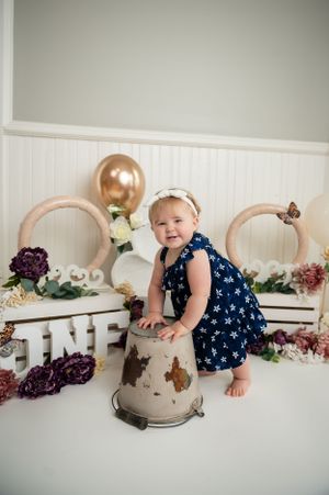 Baby in navy dress plays on white backdrop with floral decorations and rose gold balloons.