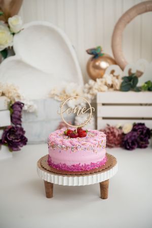 Pink birthday cake with gold topper displayed on wooden stand with floral decorations.