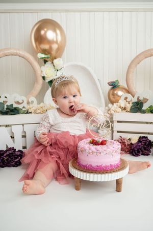 Baby in pink tulle dress reaches for decorated pink birthday cake during cake smash photo session.