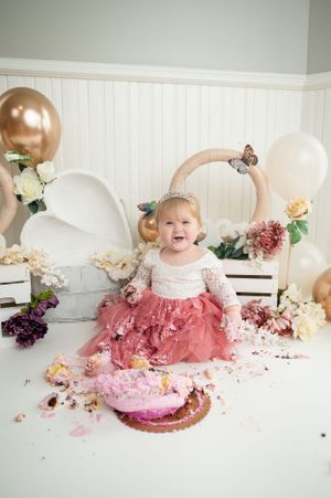 Baby laughs during cake smash session with destroyed pink cake and decorations.