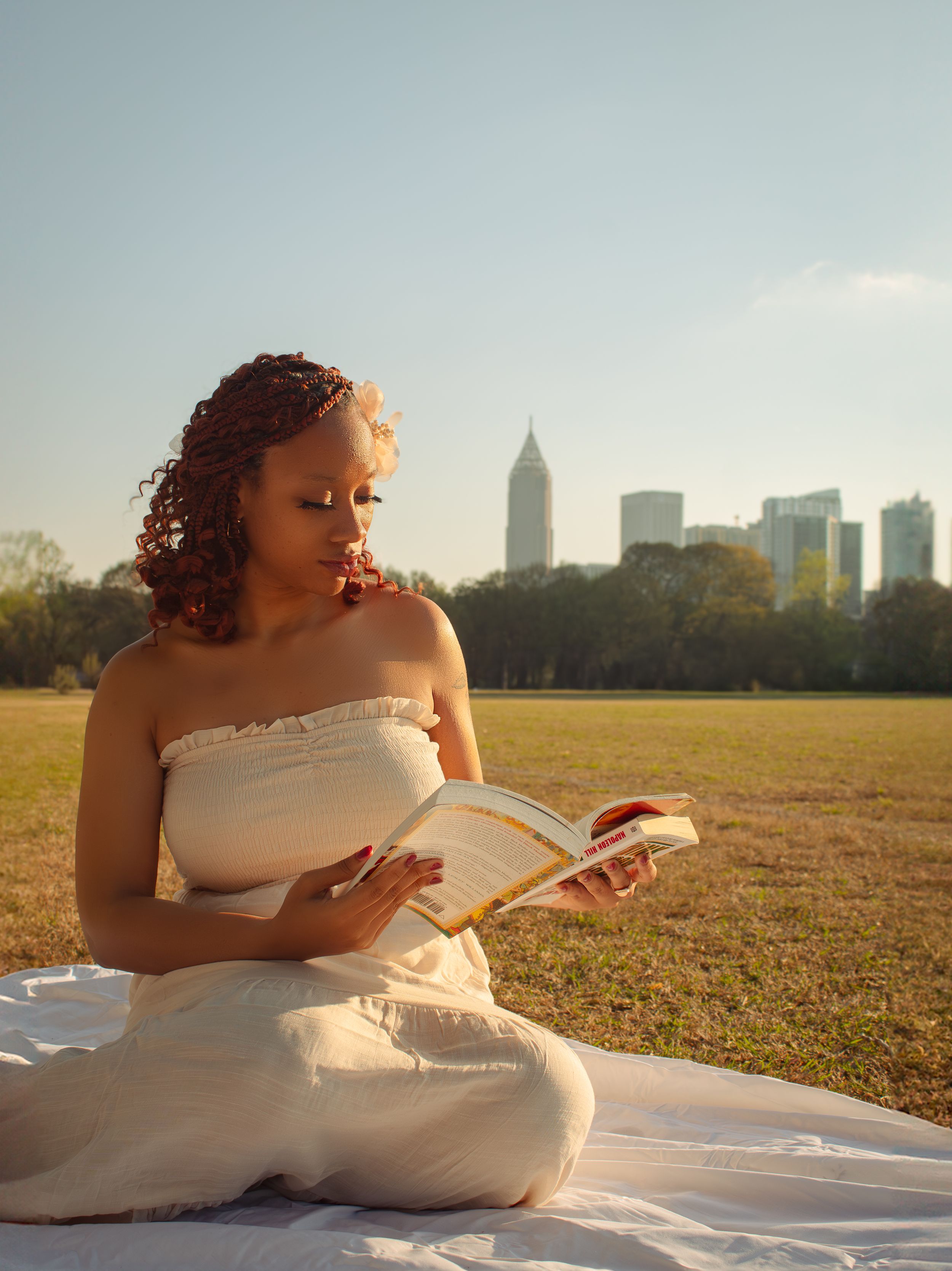 Woman in white dress reading book in park with city skyline backdrop.